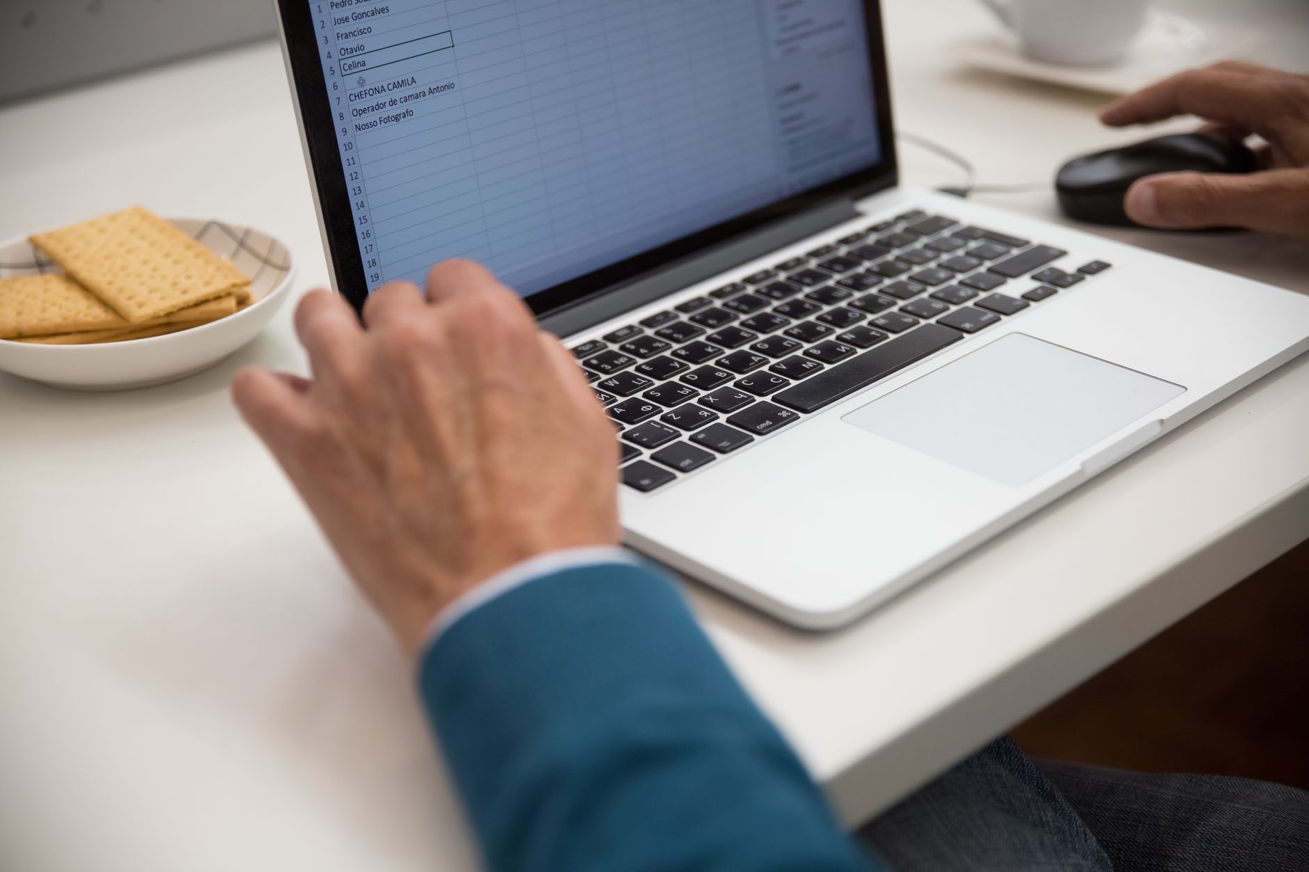 En hånd på en bærbar computer med et regneark ved siden af en skål kiks